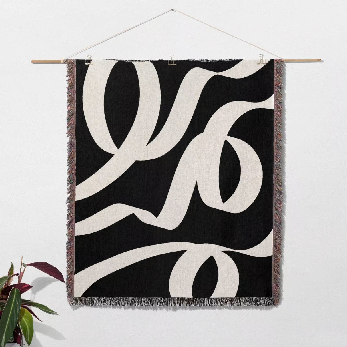 Black and white abstract waves accent woven blanket throw hanging on a wall with a plant in the corner.