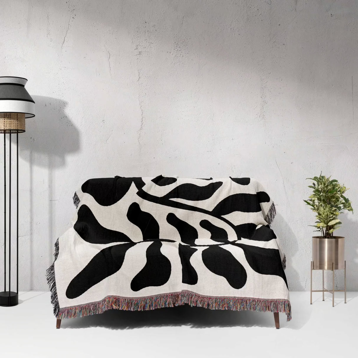 Black leaf Matisse throw blanket on a sofa against a white wall with a lamp and plant in the background.