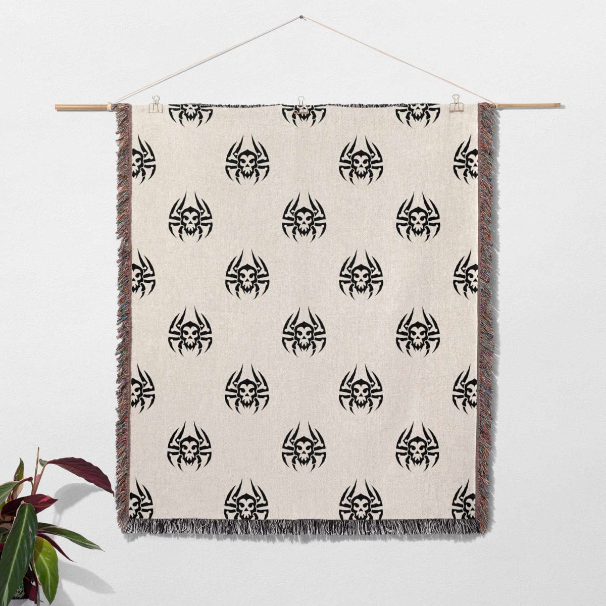 Woven blanket with black spider pattern on a beige background, hanging against a white wall.