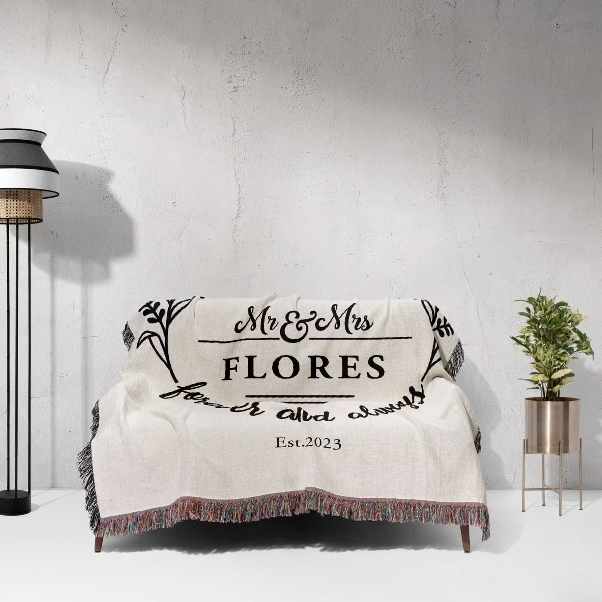 Cotton wedding anniversary gift for him woven throw blanket with 'Mr & Mrs Flores' text draped over a couch against a grey wall.