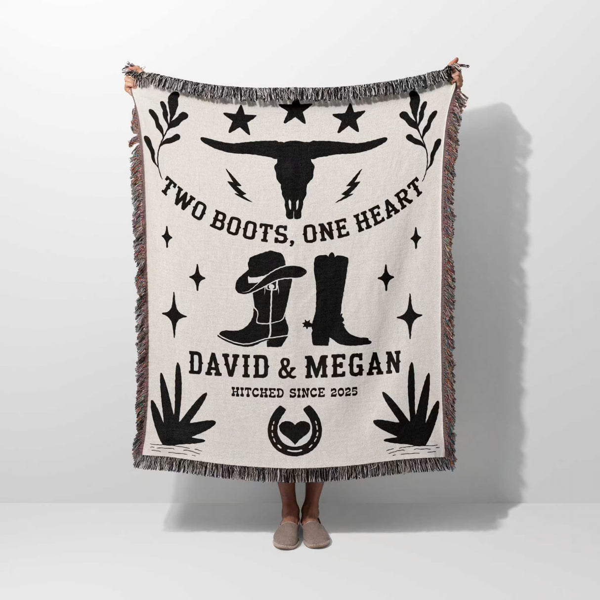 Cow skull and boots cotton anniversary gift woven throw blanket blanket with cowboy design and text on a white background held by a person.