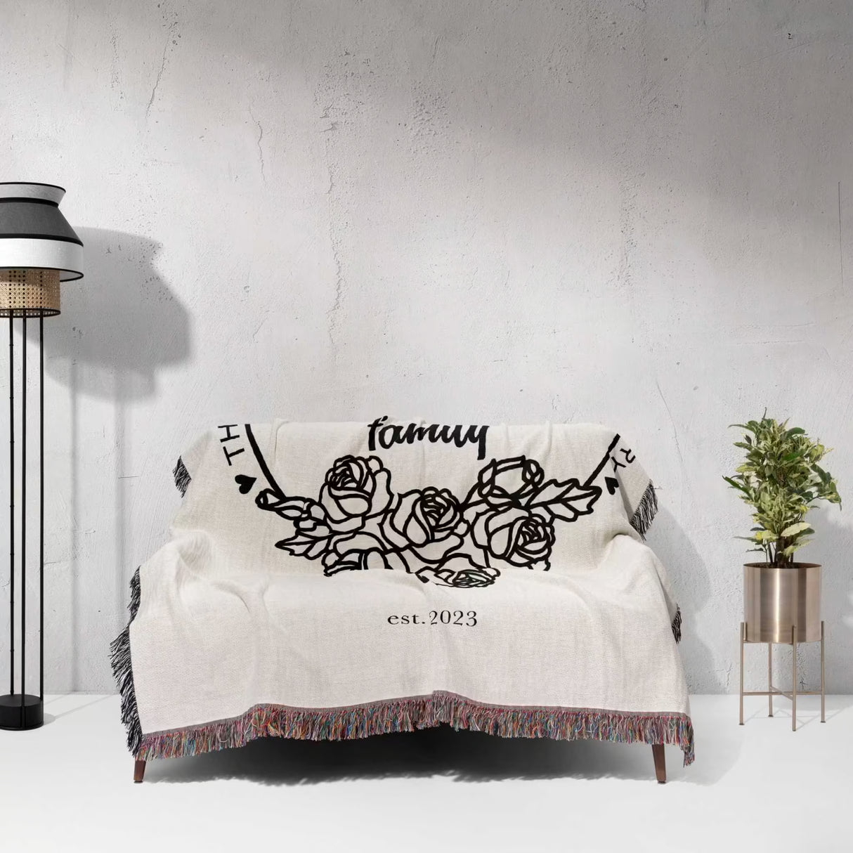 Family name personalized woven throw blanket gift for spouse on a couch featuring floral designs and text, against a light gray wall.