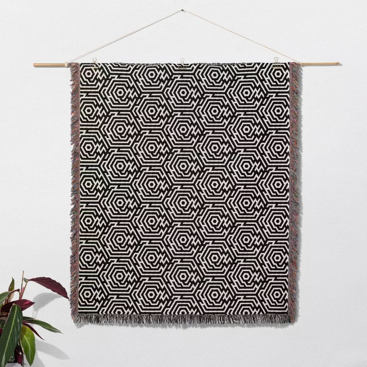 Black and white labyrinth hexagon pattern woven blanket throw handing on a white wall with a plant to the left.
