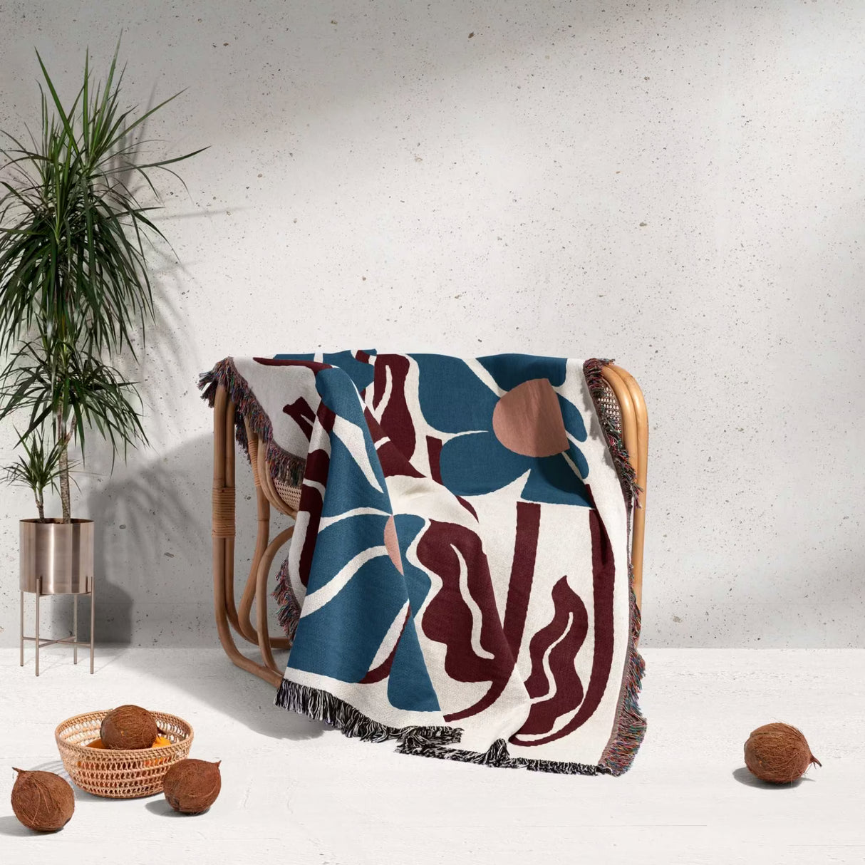 Matisse inspired blue and red tapestry throw blanket for couch with a floral pattern draped over a chair, with a plant and coconuts in the background.