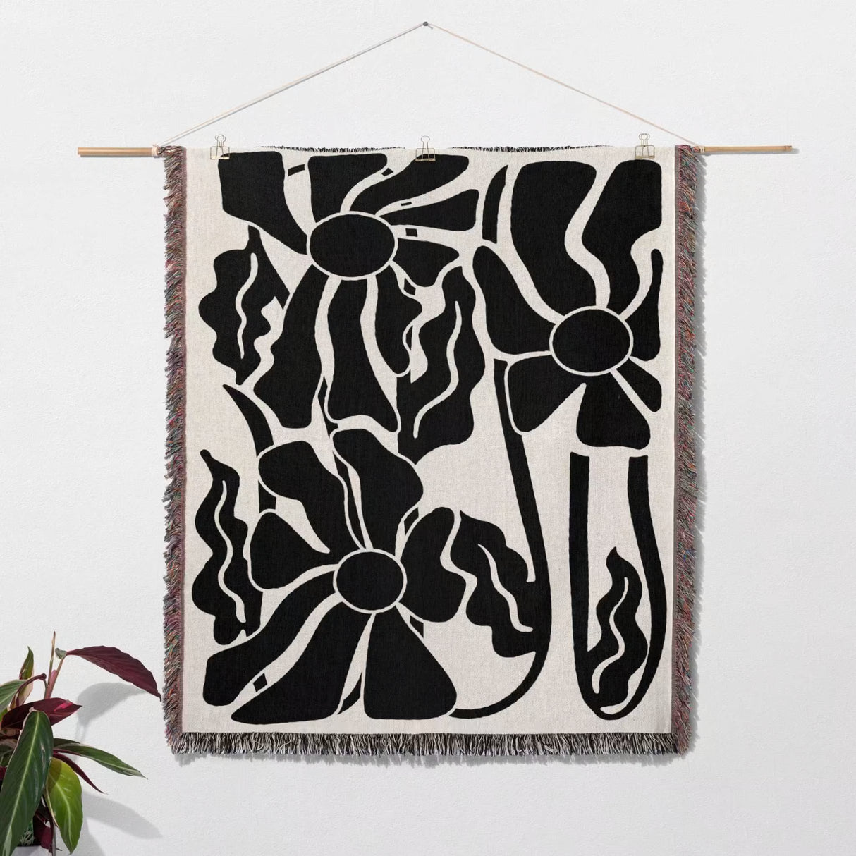 Matisse style black and white botanical cotton woven throw blanket tapestry hanging on a white wall with a plant in the corner.