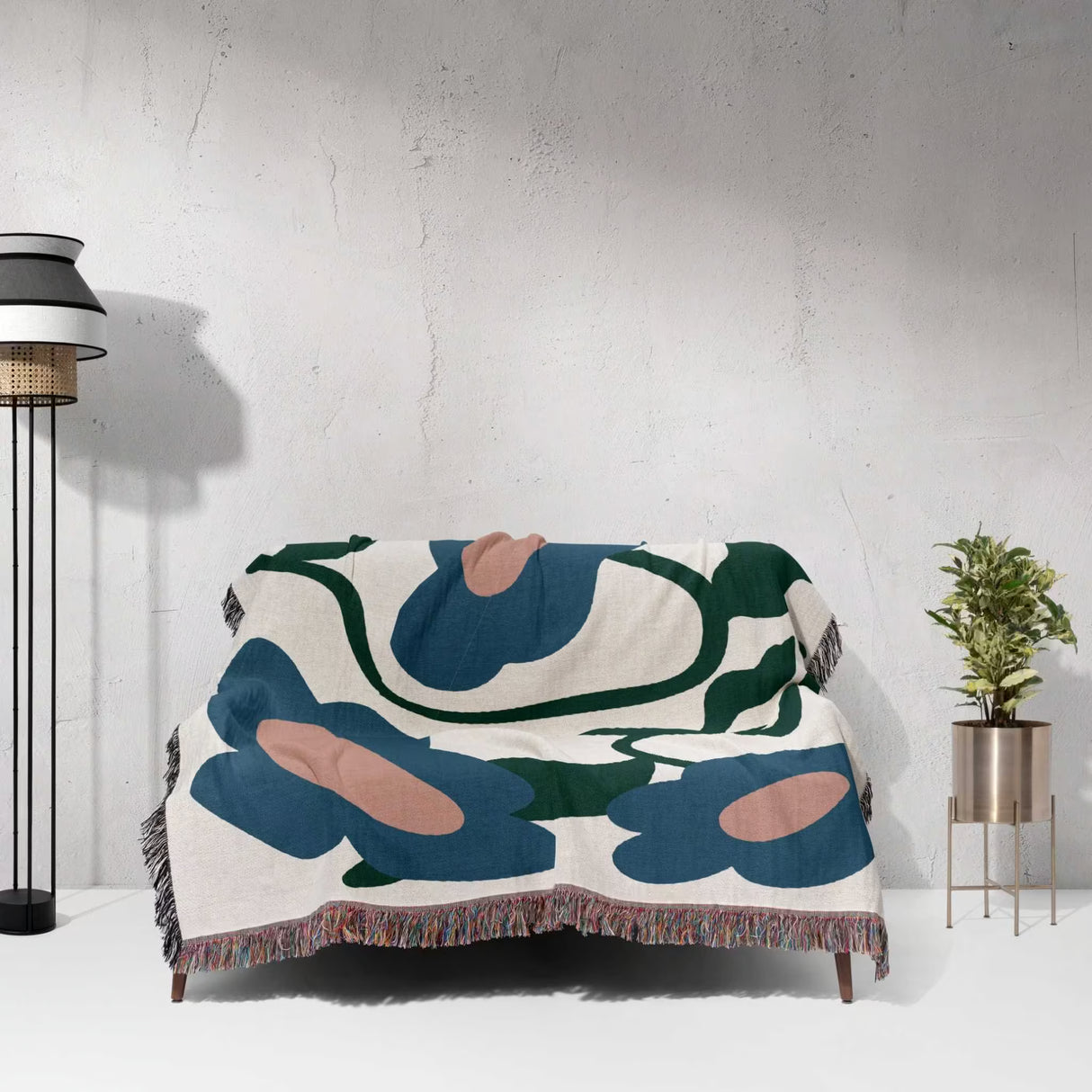 Abstract floral Matisse style flowers cotton woven throw blanket draped over a couch against a white wall with a lamp and plant in the background.