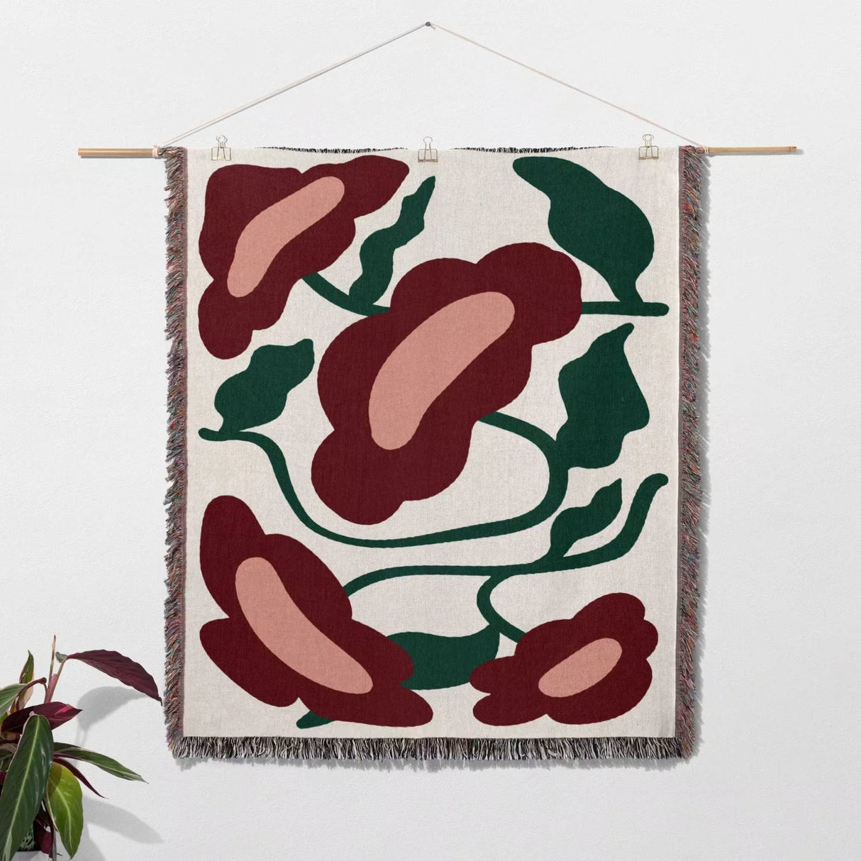 Floral patterned Matisse style red flowers cotton woven throw blanket as tapestry hanging on a white wall with a plant on the side.