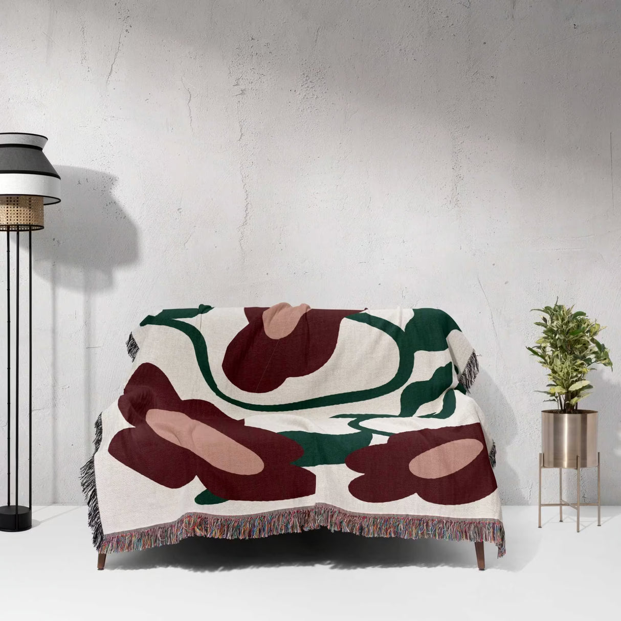 Abstract patterned Matisse style red flowers cotton woven throw blanket draped over a chair against a white wall with a lamp and plant in the background.