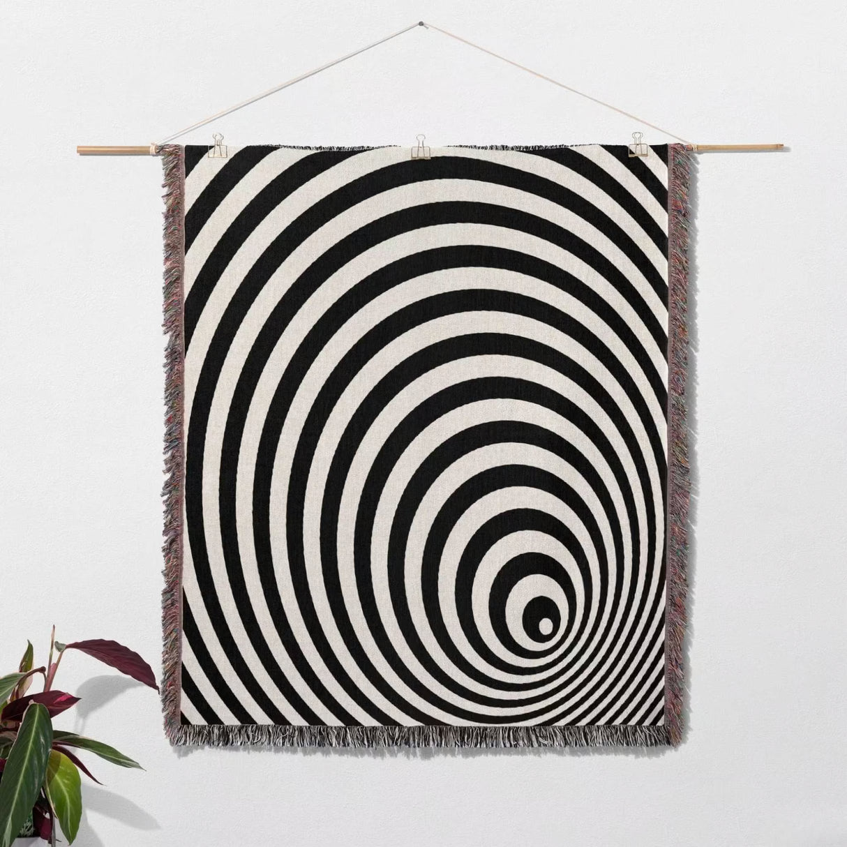Black and white optical illusion wormhole woven blanket throw tapestry hanging on a white wall with a plant in the corner.
