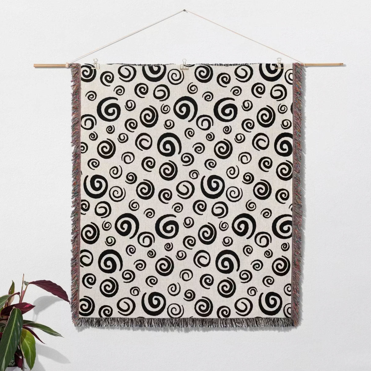 Black and white swirls patterned woven blanket throw with fringe hanging on a wooden rod against a white wall.