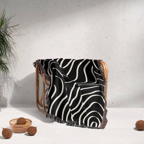 Black and white abstract art waves cotton woven blanket throw tapestry draped over a wooden chair against a light gray wall.
