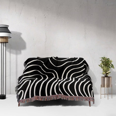 Black and white abstract art waves cotton woven blanket throw tapestry draped over a couch against a gray wall.