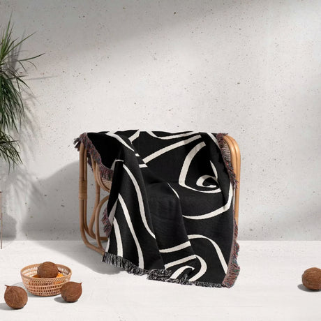Black and white abstract art waves decorative woven blanket throw draped over a wooden chair against a light gray wall.