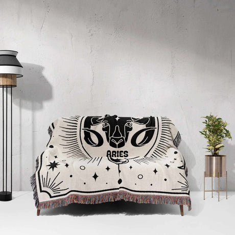 Showcasing the Aries Zodiac Blanket spread over a couch, this image highlights its decorative potential in a living space. The black and white woven design and vibrant fringe make it a unique and eye-catching birthday gift idea for both men and women.