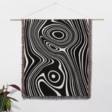 Black and white abstract woven throw blanket tapestry hanging on a white wall with a plant in the corner.
