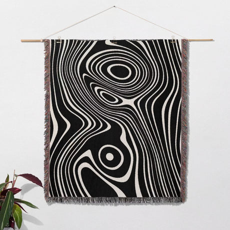 Black and white abstract woven throw blanket tapestry hanging on a white wall with a plant in the corner.