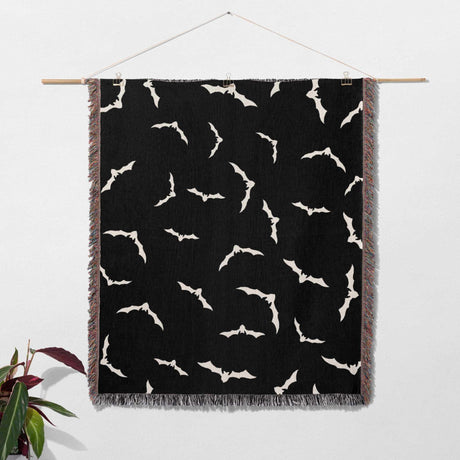 The blanket is hung on a wall like a tapestry, displaying the intricate bat pattern. The multicolored fringe adds a decorative border, making it a unique piece of wall art for Halloween.