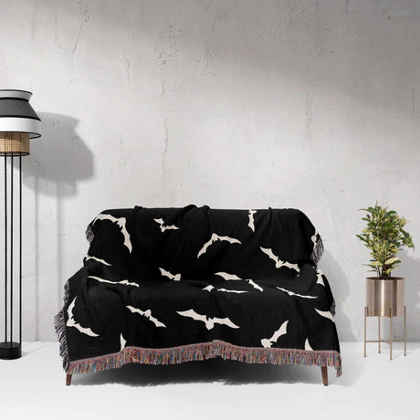 The blanket is casually draped over a couch, adding a festive Halloween touch to the living room. The white bats stand out against the black fabric, with the multicolored fringe adding a colorful accent.