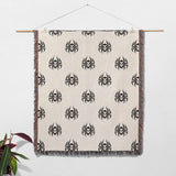 Woven blanket with black spider pattern on a beige background, hanging against a white wall.