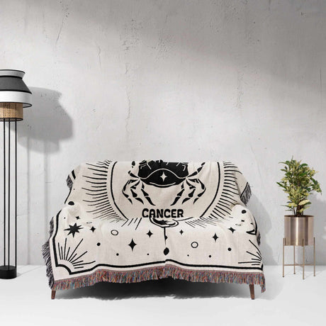 This image shows a Cancer Zodiac Blanket draped over a couch. The woven tapestry blanket features a bold crab design with celestial motifs, enhancing living spaces with astrological charm and comfort. It makes a unique birthday gift idea for both women and men.