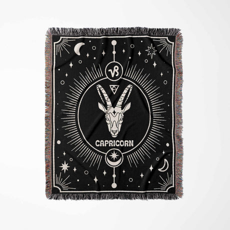 A vertically laid out Capricorn Zodiac Blanket displaying the intricate design of the Capricorn symbol at the center. The black background contrasts with white woven details and celestial elements, bordered by a multicolored fringe.