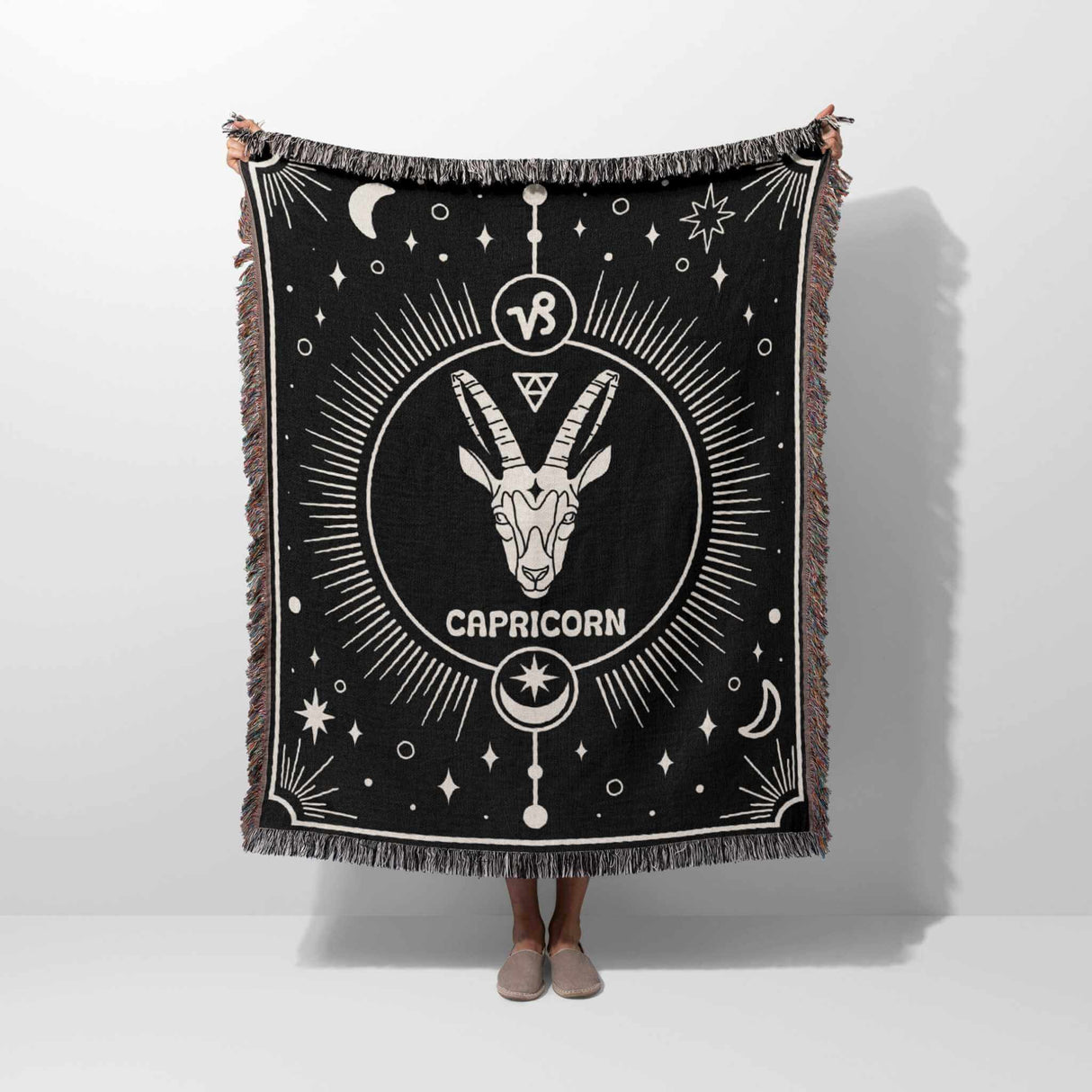 A person holding up the Capricorn Zodiac Blanket vertically, fully displaying the intricate Capricorn symbol at the center and celestial motifs against a black background. The multicolored fringe frames the entire blanket.