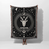 A person holding up the Capricorn Zodiac Blanket vertically, fully displaying the intricate Capricorn symbol at the center and celestial motifs against a black background. The multicolored fringe frames the entire blanket.