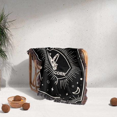 A cozy Capricorn Zodiac Blanket draped over a wicker chair. The blanket's detailed design is partially visible, showcasing the Capricorn symbol and celestial motifs against a black background. The multicolored fringe adds a decorative touch to the chair.