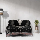 The Capricorn Zodiac Blanket elegantly spread over a couch, displaying the Capricorn symbol in the center with surrounding celestial designs. The black and white woven details stand out, complemented by the multicolored fringe at the edges.