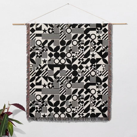 Black and white classical checker board pattern woven blanket throw hanging on a white wall with a plant in the corner.