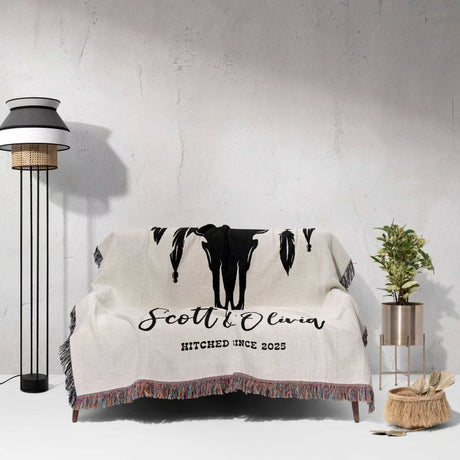 Cow skull wedding cotton anniversary gift woven throw blanket with custom 'Scott & Olivia Hitched Since 2025' text on a couch against a white wall.