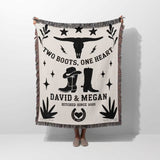 Cow skull and boots cotton anniversary gift woven throw blanket blanket with cowboy design and text on a white background held by a person. 