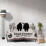 Cowboy and cowgirl western cotton anniversary gift woven throw blanket with black silhouettes of a couple and text on a white couch against a light gray wall.