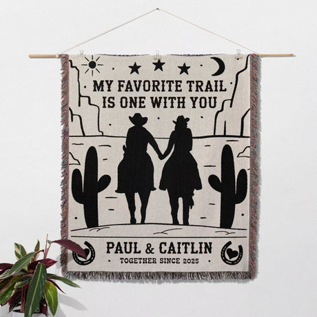 Cowboy and cowgirl on two horses cotton anniversary gift woven throw blanket with desert scene, couple holding hands, and text 'My favorite trail is one with you' by Paul & Caitlin.
