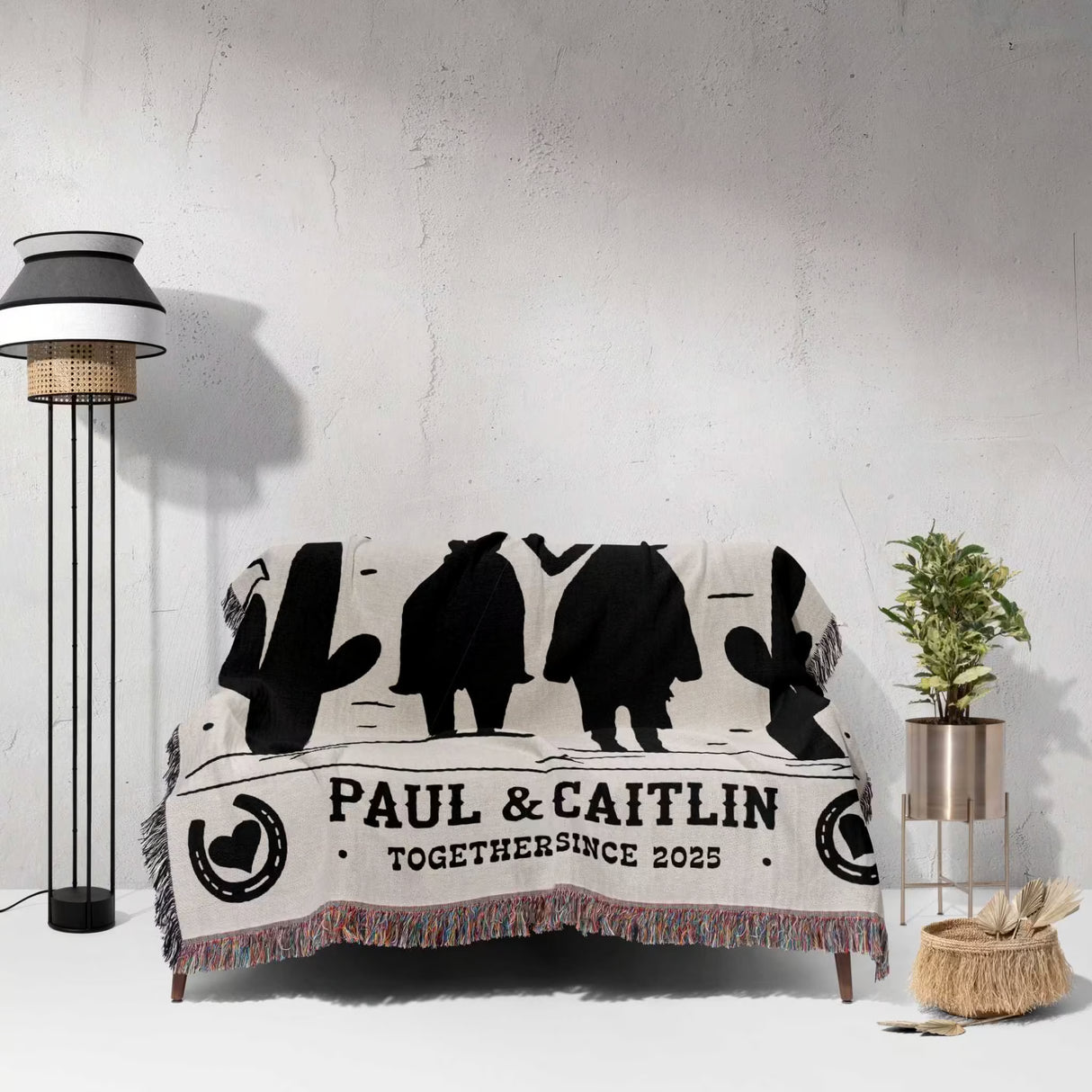 Cowboy and cowgirl on two horses cotton anniversary gift woven throw blanket with black silhouettes and text draped over a couch in a room with a lamp and plant.