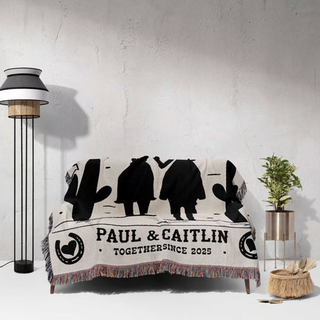 Cowboy and cowgirl on two horses cotton anniversary gift woven throw blanket with black silhouettes and text draped over a couch in a room with a lamp and plant.