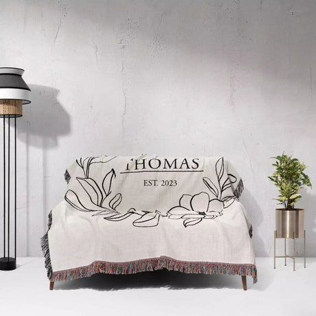 Custom family name cotton throw blanket with floral design and 'THOMAS EST. 2023' text draped over a chair against a white wall.