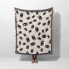 A person is holding up the Cute Halloween Blanket to show its full size and design. The blanket’s playful pattern of scattered black spiders against an off-white backdrop is fully visible, with the multicolored fringe neatly bordering the edges.