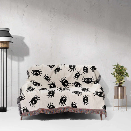 This image showcases the Cute Halloween Blanket spread out over a couch, demonstrating its perfect fit for adding a touch of Halloween charm to any seating area. The blanket’s design, featuring cute black spiders on an off-white background, paired with the vibrant multicolored fringe, makes it an ideal decorative and functional piece.