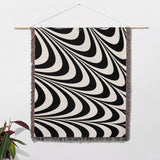 Black and white decorative abstract waves woven blanket throw tapestry with fringe hanging on a wooden rod against a white wall.