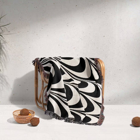 Black and white decorative abstract waves woven blanket throw tapestry draped over a wooden stool against a light gray background.