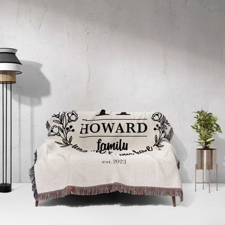 Family monogrammed woven throw blanket for couch and bed with 'HOWARD family est. 2023' text draped over a couch against a gray wall.