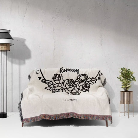 Family name personalized woven throw blanket gift for spouse on a couch featuring floral designs and text, against a light gray wall.