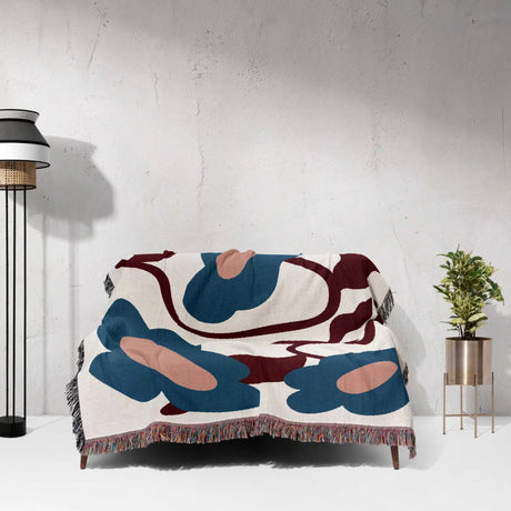 Floral Matisse inspired tapestry throw blanket for couch draped over a couch against a white wall with a lamp and plant in the background.