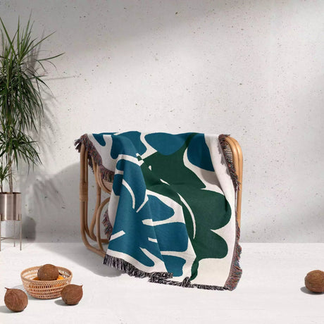 Blue and green leaf-patterned Henri Matisse blue flowers woven throw blanket draped over a wooden chair against a light gray wall.