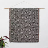 Black and white labyrinth hexagon pattern woven blanket throw handing on a white wall with a plant to the left.