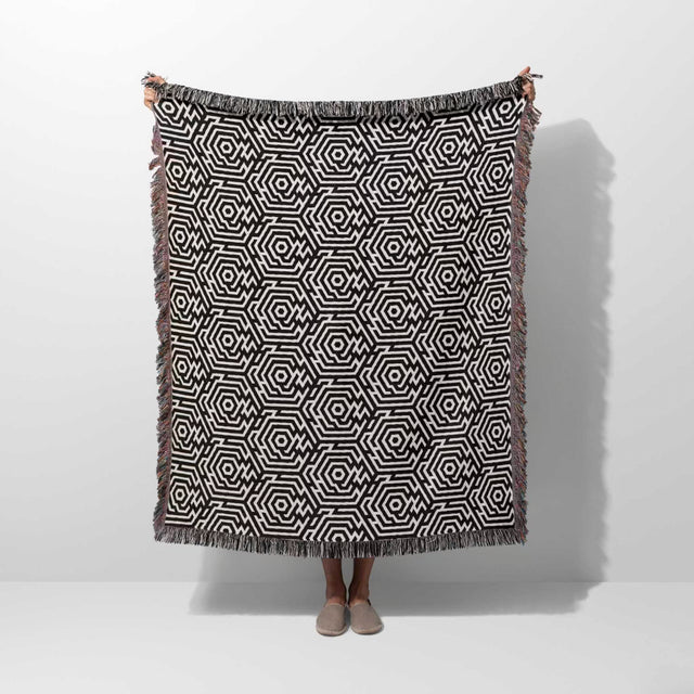 Black and white labyrinth hexagon pattern woven blanket throw held by a person on a white background.