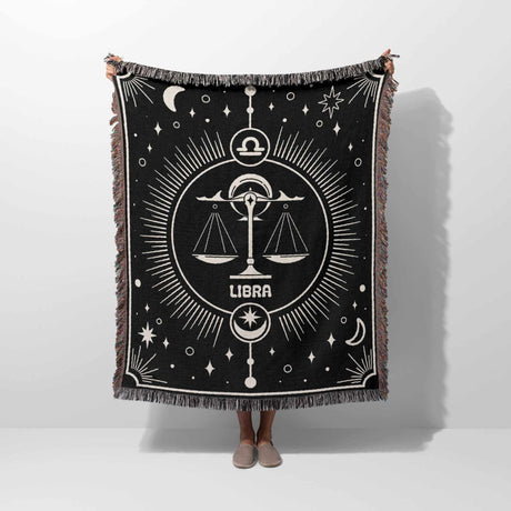 The Libra Zodiac Blanket held up by a person, showing its full vertical design. The scales symbol is prominently displayed in the center, with celestial decorations around it, and the multicolor fringe completing the look.