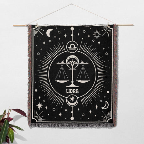 The Libra Zodiac Blanket hung on a wall as a piece of art, emphasizing its decorative potential beyond just being a blanket. The central scales symbol, celestial elements, and multicolor fringe create a striking visual display.