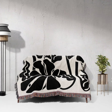 Matisse style black and white botanical cotton woven throw blanket on a couch with a lamp and plant. 